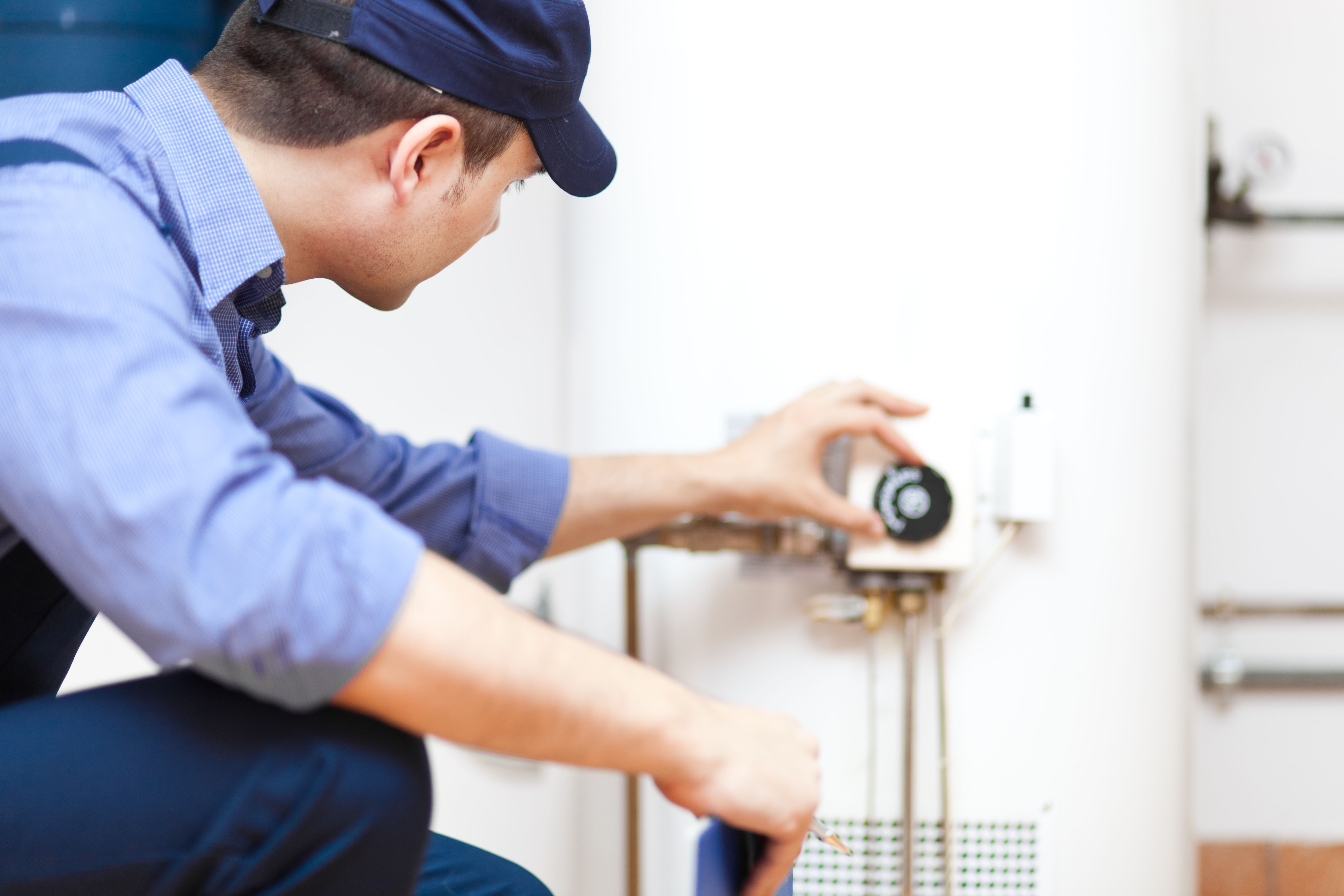 A plumber wearing a blue shirt and hat is adjusting the thermostat on a water heater